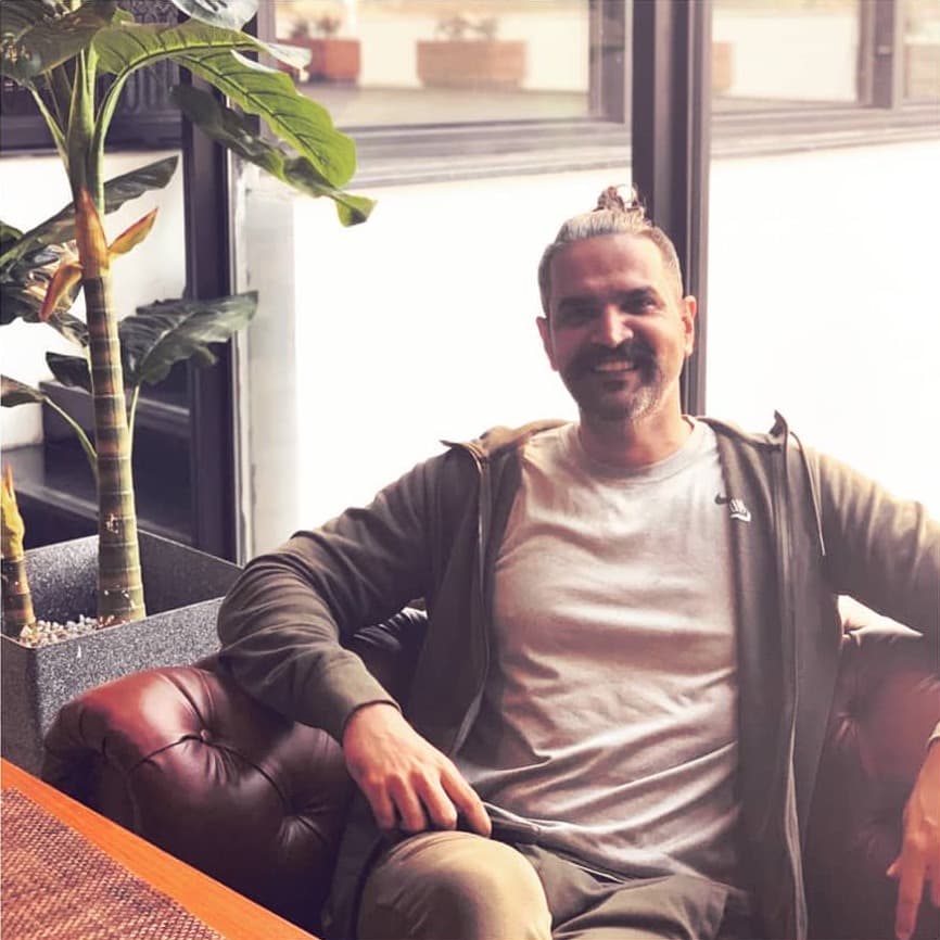 Farrukh Hammad smiling, seated on a leather sofa in soft window light beside a tall plant